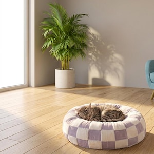 May include: A round, plush pet bed featuring a checkered pattern in soft lavender and white. A brown tabby cat is curled up asleep in the center of the bed, which is placed on a light-colored wooden floor in a sunlit room.