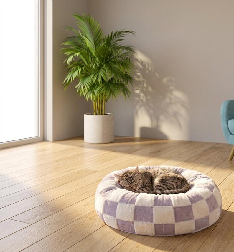 May include: A round, plush pet bed featuring a checkered pattern in soft lavender and white. A brown tabby cat is curled up asleep in the center of the bed, which is placed on a light-colored wooden floor in a sunlit room.