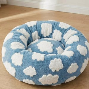 May include: A round, plush pet bed featuring a soft, textured blue fabric base with a repeating pattern of white, fluffy cloud shapes. The bed has a raised, cushioned rim and a central circular cushion, providing a cozy, circular resting area.