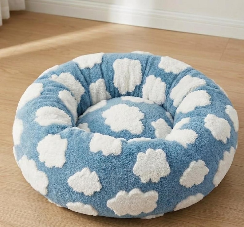 May include: A round, plush pet bed featuring a soft, textured blue fabric base with a repeating pattern of white, fluffy cloud shapes. The bed has a raised, cushioned rim and a central circular cushion, providing a cozy, circular resting area.