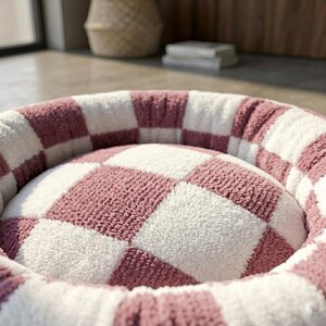 May include: A round pet bed featuring a plush, textured checkerboard pattern in alternating dusty rose pink and cream white squares. The bed has a raised, cushioned rim and a soft, padded center, constructed from a fuzzy, fleece-like material.