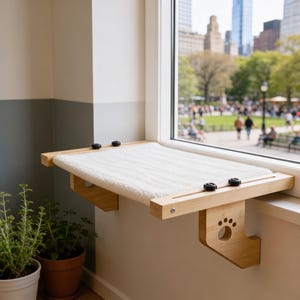 Peut inclure: Un perchoir de fenêtre en bois clair pour animaux avec une surface en tissu blanc texturé et doux. Le cadre comprend des boutons noirs réglables et des supports latéraux avec une découpe en forme de patte, conçu pour se fixer au rebord.
