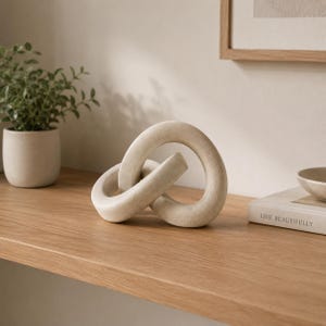 Peut inclure: Un objet sculptural beige et texturé en forme de nœud entrelacé. La pièce présente une forme arrondie et lisse avec une finition mate, créant une structure en boucle continue qui repose sur une surface plane comme objet décoratif.