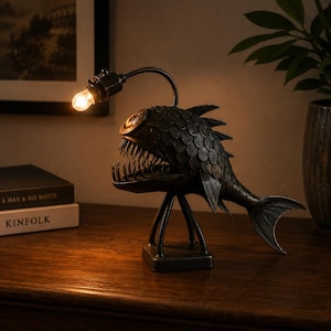 Pode incluir: Um candeeiro de mesa em metal escuro com a forma de um peixe-pescador, com escamas texturizadas, dentes afiados e uma lâmpada acesa suspensa num braço metálico curvo. O candeeiro assenta numa base quadrada com quatro pernas finas.