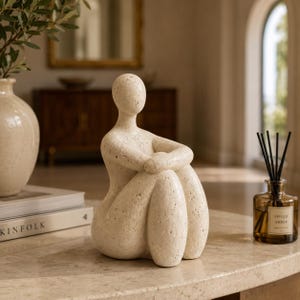 May include: A beige, textured stone sculpture of a seated figure with a smooth, rounded head and arms wrapped around its knees. The figure has a porous, speckled surface and is positioned in a curled, seated pose on a flat, light-coloured surface.