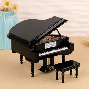 Pode incluir: Um piano de cauda em miniatura preto com acabamento brilhante, apresentando uma tampa levantada, teclas brancas e pretas, cordas internas douradas e uma pequena partitura retangular. Um banco preto a condizer com quatro pernas está à frente.