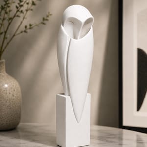 May include: A matte white sculpture of an owl with a smooth, elongated body and a rounded head, perched atop a rectangular base. The owl features a stylized, tapered design with a pointed lower section and a recessed face with closed eyes.