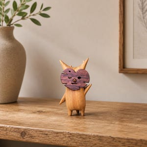 Puede incluir: Una pequeña figura de madera tallada a mano con un cuerpo de color marrón claro y textura lisa, dos orejas puntiagudas y patas cortas. Una máscara de madera en forma de corazón de color marrón oscuro con detalles recortados en ojos y boca.