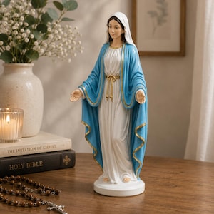 May include: A standing statue of the Virgin Mary wearing a white gown and a blue mantle with gold trim. The figure has a white head covering, a gold-colored waist tie, and arms extended outward with palms facing up, all set on a circular base.