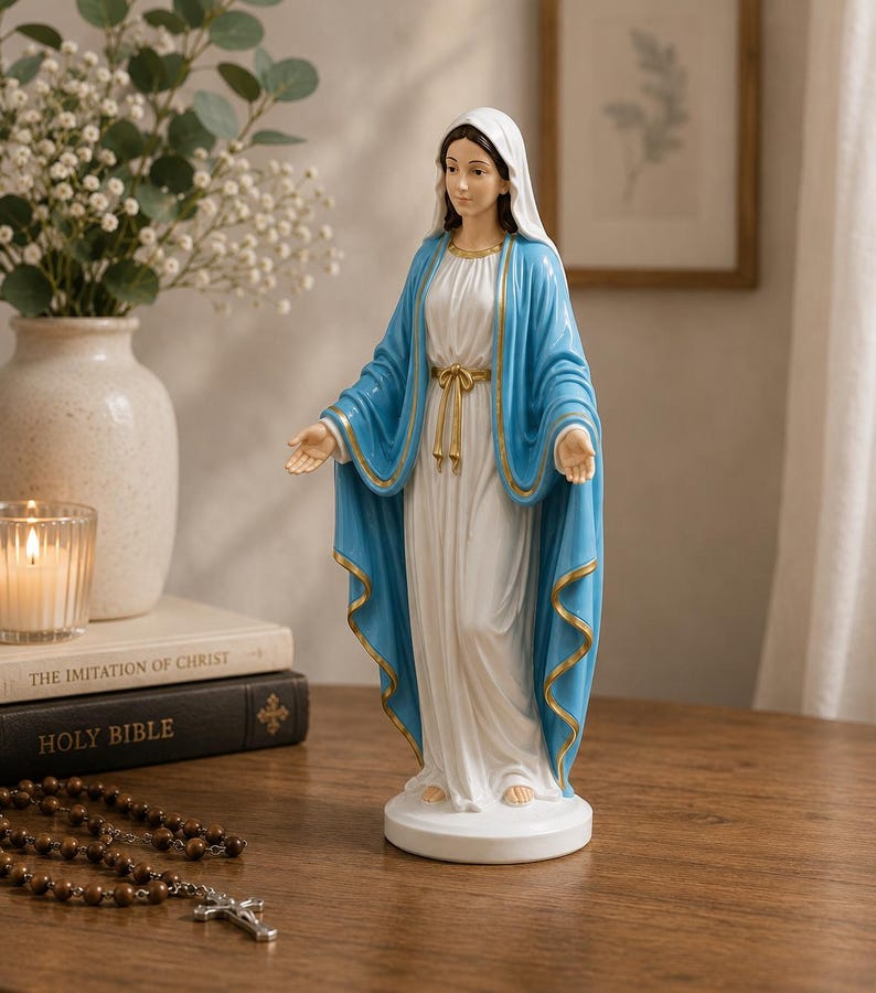 May include: A standing statue of the Virgin Mary wearing a white gown and a blue mantle with gold trim. The figure has a white head covering, a gold-colored waist tie, and arms extended outward with palms facing up, all set on a circular base.