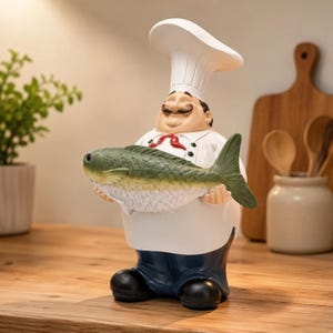 May include: A decorative chef figurine wearing a tall white hat, a white jacket with a red necktie, and dark blue trousers. The chef has a mustache and is holding a large, textured green and white fish with both hands in a horizontal position.