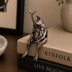 Peut inclure: Une figurine métallique argentée représentant un scarabée rhinocéros doté d'un torse humanoïde et de membres articulés. La créature est représentée en position assise, les jambes pendantes au bord d'une surface, révélant des textures détaillées.