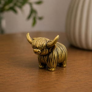 Op de afbeelding: Een klein, goudkleurig metalen beeldje van een Schotse hooglander. Het beeldje heeft een gestructureerde, ruige vacht, opvallende gebogen hoorns en een pony die de ogen bedekt, afgewerkt met een verweerde look voor extra detail.