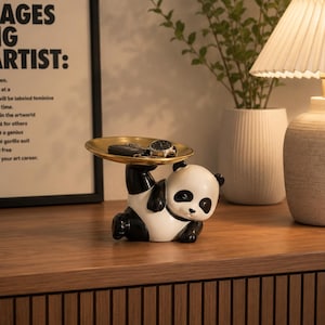 Könnte beinhalten: Eine dekorative Figur eines schwarz-weißen Pandas, der auf der Seite liegt und ein rundes, goldfarbenes Metalltablett über seinem Kopf hält. Auf dem Tablett liegen eine schwarze Armbanduhr und ein Schlüsselbund. Der Panda hat schwarze Ohren.