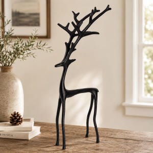 Puede incluir: Una escultura de metal negro mate de un ciervo de pie con patas alargadas y cuello esbelto. El ciervo presenta astas ramificadas prominentes que se extienden hacia arriba y hacia afuera, creando una silueta alta y estilizada con acabado oscuro.