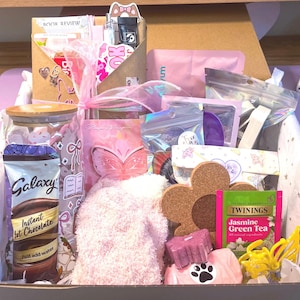May include: A gift box containing a glass tumbler with a bamboo lid, a packet of Galaxy instant hot chocolate, a pair of fuzzy pink socks, a flower-shaped cork coaster, a Twinings Jasmine Green Tea bag, a yellow butterfly clip, and various stationery items.