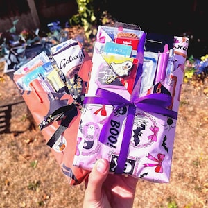 May include: Two rectangular gift packages wrapped in Halloween-themed paper. The left package is orange with black cat motifs, tied with a black ribbon. The right package is white with pink bows, bats, and the text BOO!, tied with a purple ribbon.