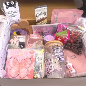 May include: A gift box containing a glass tumbler with a bamboo lid and the text Enemies to Lovers Cola, a pink butterfly notepad, a Galaxy instant chocolate packet, a cork coaster, a purple candle, a pink towel, and various small accessories.