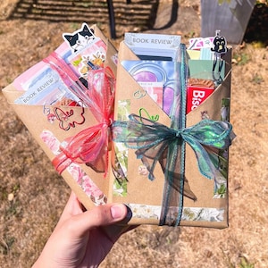 Puede incluir: Dos sobres de papel marrón con artículos de papelería, cada uno atado con una cinta transparente. El sobre izquierdo tiene una cinta rosa y una pegatina de gato, el derecho una cinta verde azulado y un clip de gato. Ambos incluyen el texto BOOK REVIEW.