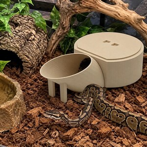 May include: A beige, 3D-printed reptile hide with a circular base and an attached, elevated bowl-like entrance. The structure features a flat top with two small square cutouts and is positioned on a substrate of wood chips near a snake.