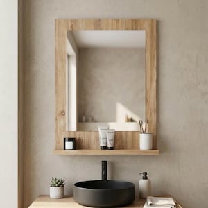 May include: A rectangular wall mirror with a light brown wood grain frame and an integrated shelf at the bottom. The shelf holds a black candle, two white tubes of toiletries, and a white cup containing two wooden toothbrushes.