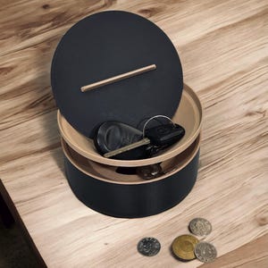 May include: A round, matte black storage container with a light-coloured wooden rim and a matching circular lid featuring a thin wooden handle. The container holds a set of black keys and a metal ring, while three metallic coins rest nearby.