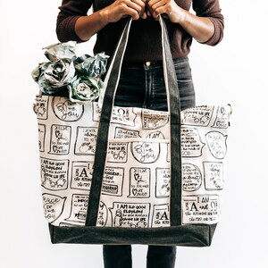 May include: A large, white canvas tote bag with black straps and a black canvas bottom. The bag is printed with black and white text and illustrations of sheep, hearts, and other religious symbols. The text includes phrases like "I am the good shepherd" and "I love you".