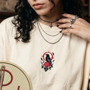 May include: An off-white t-shirt featuring a small, centred embroidery of a black raven perched above a red rose, framed by a red circular line and thorns. A circular patch in the bottom left corner displays a needle and thread with the text EMBROIDERY.