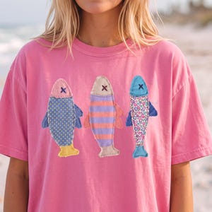 Puede incluir: Una camiseta de manga corta de color rosa brillante con un gráfico de tres peces dispuestos uno al lado del otro. Cada pez tiene un patrón distinto: uno con lunares amarillos, otro con rayas horizontales naranjas y moradas, y otro con estampado floral.