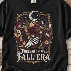 May include: A black short-sleeve t-shirt featuring a graphic of a skeleton hand holding a bouquet of autumn flowers, leaves, and a crescent moon. The text "FOREVER IN MY FALL ERA" is printed in a decorative font below the floral illustration.