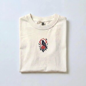 May include: A cream-coloured short-sleeve t-shirt featuring a central embroidered graphic of a black raven perched on a red rose. The design includes a red circular frame, small stars, and a diamond shape with the letter S at the bottom.