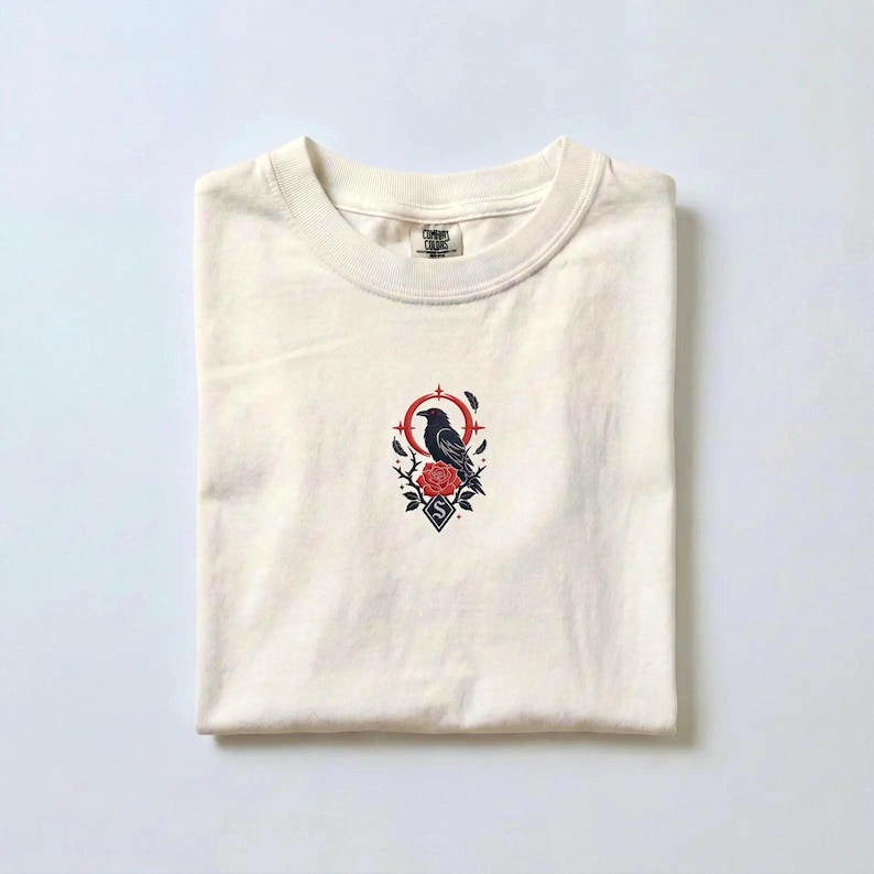 May include: A cream-coloured short-sleeve t-shirt featuring a central embroidered graphic of a black raven perched on a red rose. The design includes a red circular frame, small stars, and a diamond shape with the letter S at the bottom.