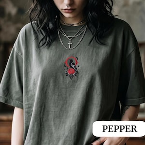 May include: A dark olive green t-shirt featuring a central embroidered design of a black crow perched above a red rose, framed by a thin red circle and black leaves. The shirt includes a small diamond-shaped emblem with the letter S below the rose.