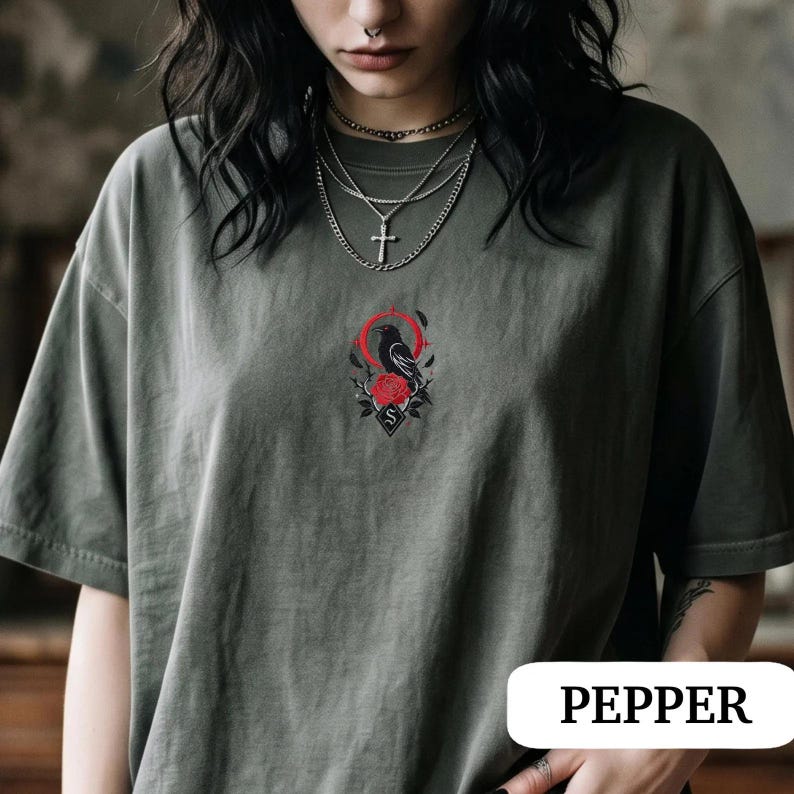 May include: A dark olive green t-shirt featuring a central embroidered design of a black crow perched above a red rose, framed by a thin red circle and black leaves. The shirt includes a small diamond-shaped emblem with the letter S below the rose.