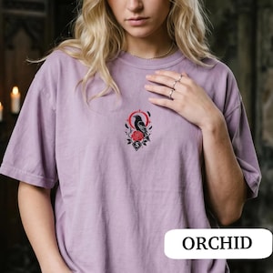 May include: A light purple short-sleeve t-shirt featuring a central embroidered graphic of a black raven perched above a red rose, framed by a red circular element and small black feathers. The shirt has a crew neckline and a relaxed fit.