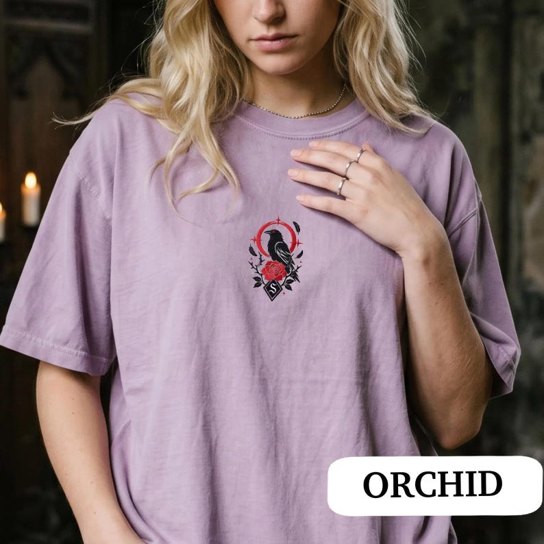 May include: A light purple short-sleeve t-shirt featuring a central embroidered graphic of a black raven perched above a red rose, framed by a red circular element and small black feathers. The shirt has a crew neckline and a relaxed fit.