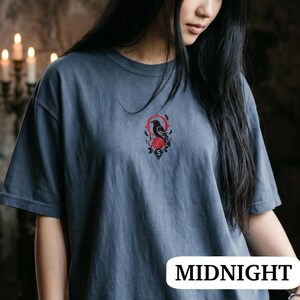 May include: A slate blue short-sleeved t-shirt featuring a small, centred graphic on the chest. The design depicts a black raven perched above a red rose, framed by a thin red circular line and a small black diamond shape containing the letter S.