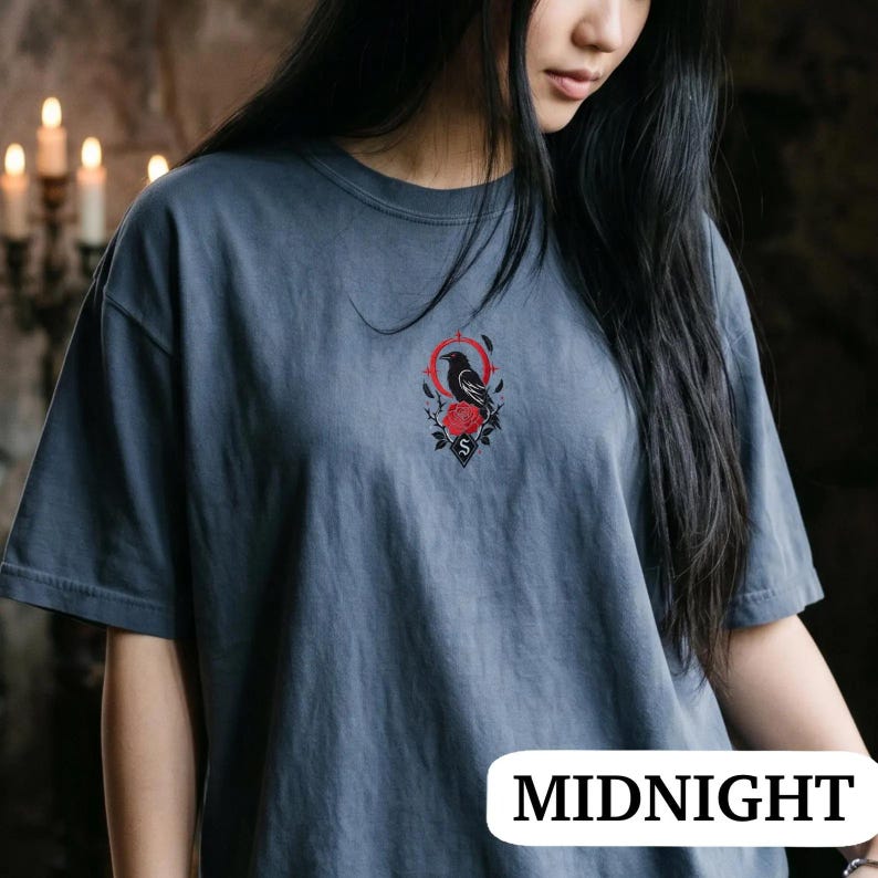 May include: A slate blue short-sleeved t-shirt featuring a small, centred graphic on the chest. The design depicts a black raven perched above a red rose, framed by a thin red circular line and a small black diamond shape containing the letter S.