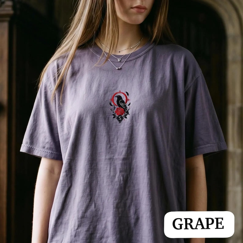 May include: A grape-coloured short-sleeve t-shirt featuring a small, centred embroidery of a black raven perched above a red rose, framed by a red circular line and a diamond shape containing the letter S, with a crew neckline and loose fit.