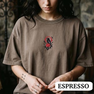May include: A brown crew neck t-shirt featuring a small, centred graphic of a black crow perched above a red rose, surrounded by a thin red circular frame and black leaf accents. The design includes a small black diamond shape with the letter S.