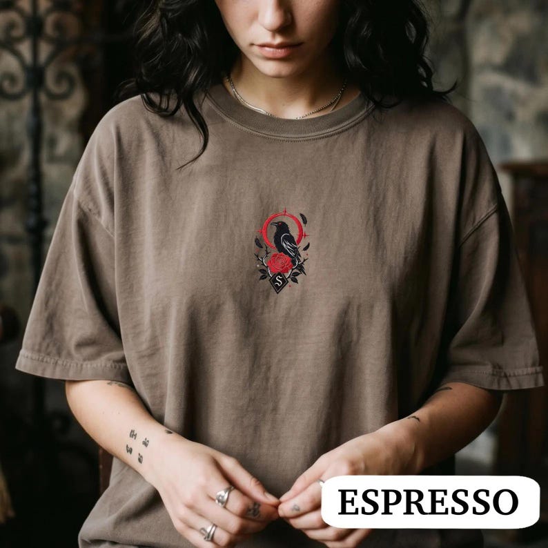 May include: A brown crew neck t-shirt featuring a small, centred graphic of a black crow perched above a red rose, surrounded by a thin red circular frame and black leaf accents. The design includes a small black diamond shape with the letter S.