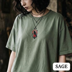 May include: A sage green short-sleeve t-shirt featuring a central embroidered graphic of a black crow perched above a red rose, framed by a red circle and a crescent moon. The design includes a small diamond shape with the letter S at the bottom.