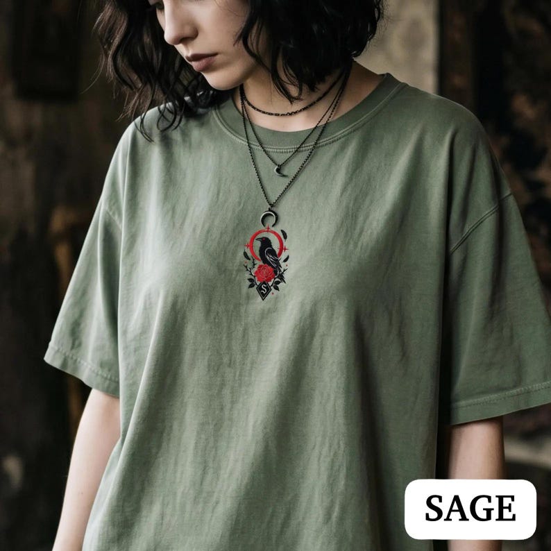 May include: A sage green short-sleeve t-shirt featuring a central embroidered graphic of a black crow perched above a red rose, framed by a red circle and a crescent moon. The design includes a small diamond shape with the letter S at the bottom.
