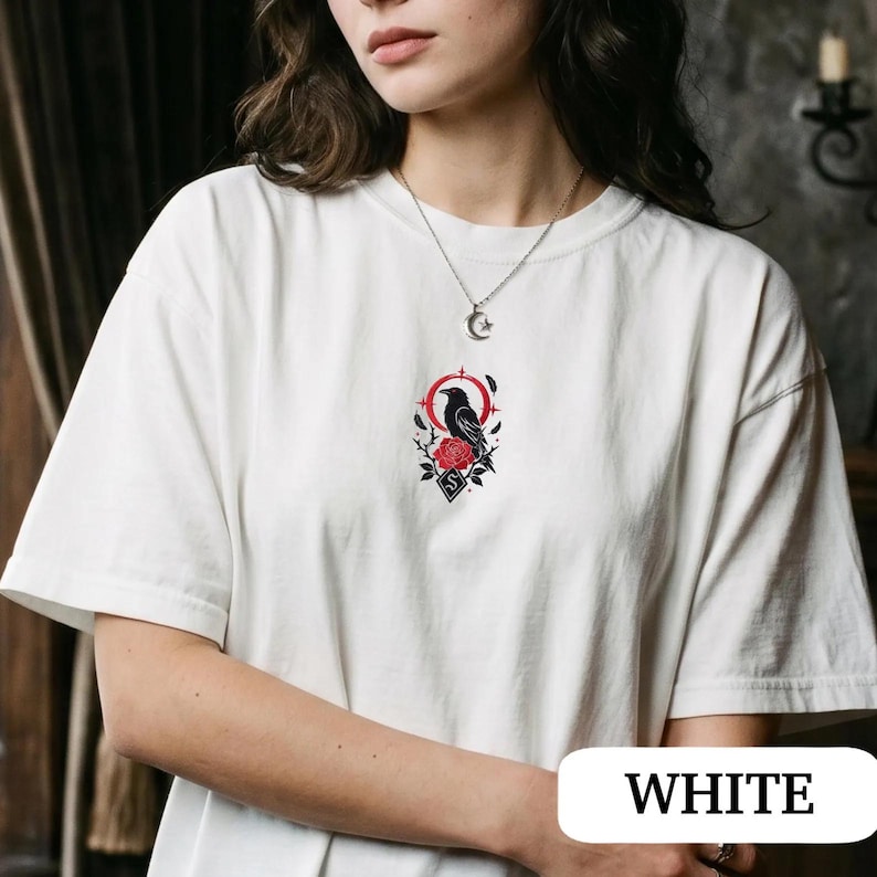 May include: A white short-sleeved t-shirt featuring a central graphic print of a black crow perched on a red rose, surrounded by a red circular frame with star accents and small black leaves, set against a plain white fabric background.