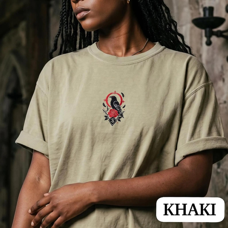 May include: A khaki t-shirt featuring a central graphic design of a black raven perched above a red rose, surrounded by a thin red circular frame and small black thorns. The design includes a small diamond shape with the letter S at the bottom.