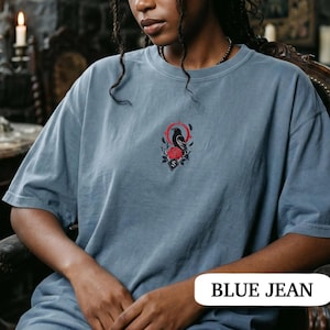 May include: A blue jean coloured t-shirt featuring a small, centred embroidery design. The graphic depicts a black raven perched above a vibrant red rose, framed by a thin red circular line and subtle black leaf accents with a small letter S.