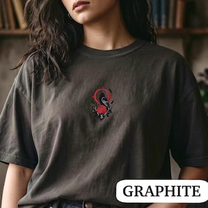 May include: A graphite grey crew neck t-shirt featuring a central embroidered graphic of a black crow perched above a red rose, framed by a thin red circle and decorative elements. The shirt has short sleeves and a relaxed, loose-fitting silhouette.