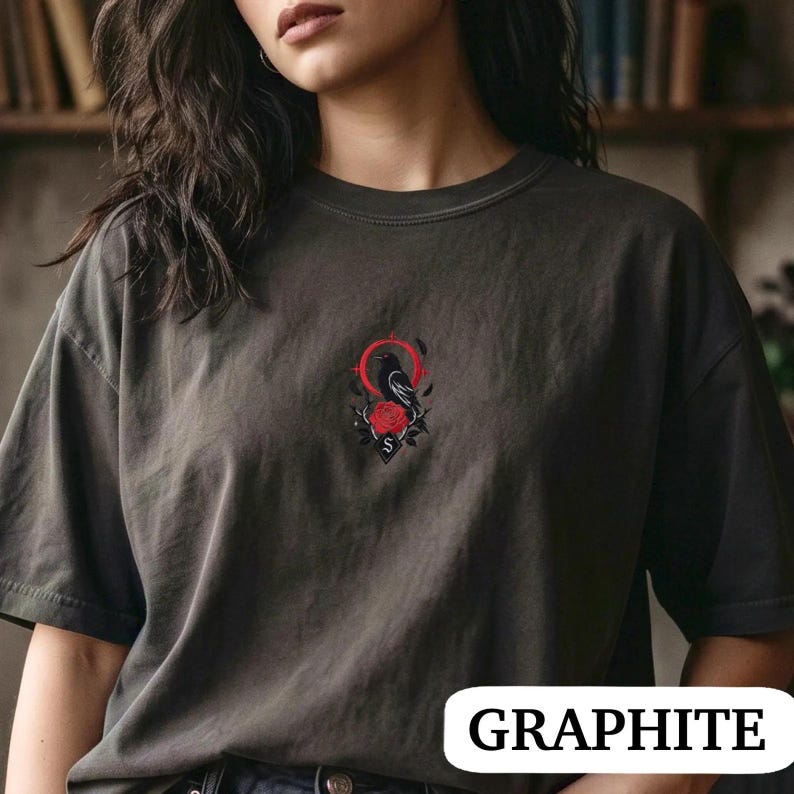May include: A graphite grey crew neck t-shirt featuring a central embroidered graphic of a black crow perched above a red rose, framed by a thin red circle and decorative elements. The shirt has short sleeves and a relaxed, loose-fitting silhouette.