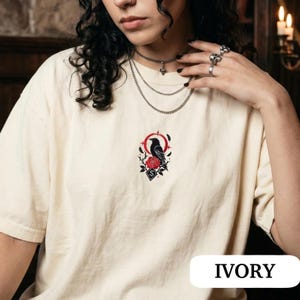 May include: An ivory t-shirt featuring a central embroidered design of a black raven perched above a red rose, surrounded by a red circular frame and thorny branches. The graphic includes a small diamond shape with a white letter S inside.