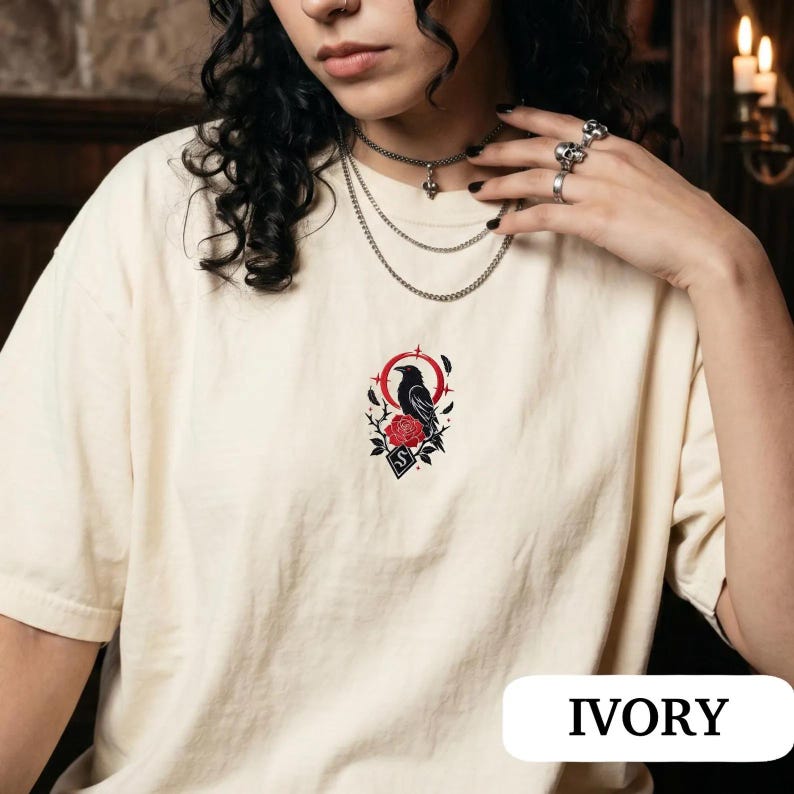 May include: An ivory t-shirt featuring a central embroidered design of a black raven perched above a red rose, surrounded by a red circular frame and thorny branches. The graphic includes a small diamond shape with a white letter S inside.